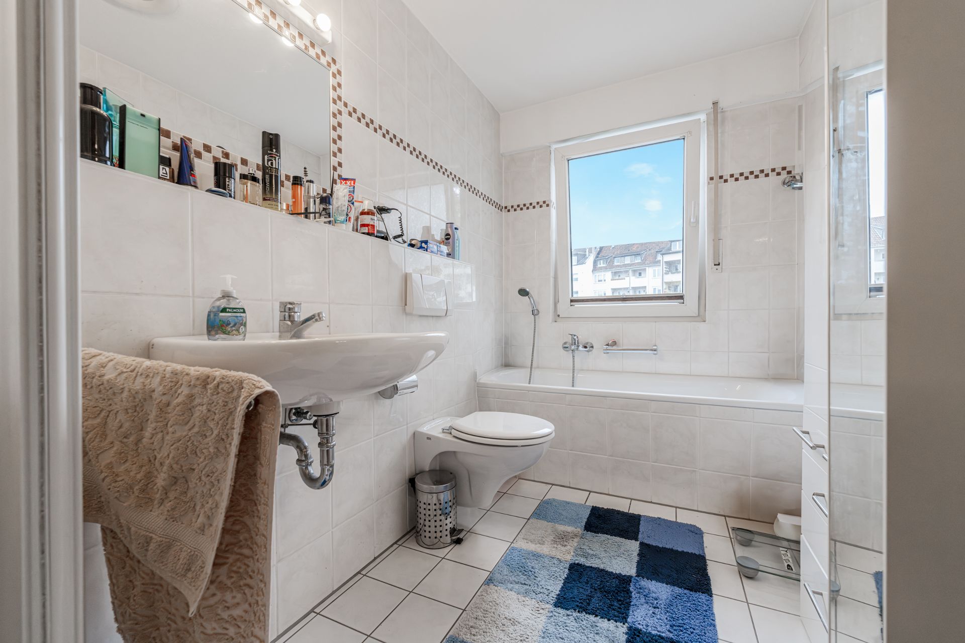 Helles Bad mit Waschbecken, WC, Badewanne, Fenster und blau kariertem Teppich.