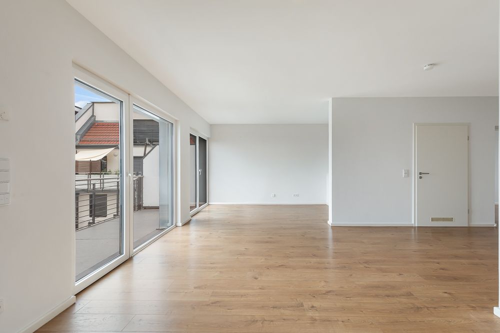Heller leerer Raum mit Holzboden, weißen Wänden und großen Fenstern zu einem Balkon.