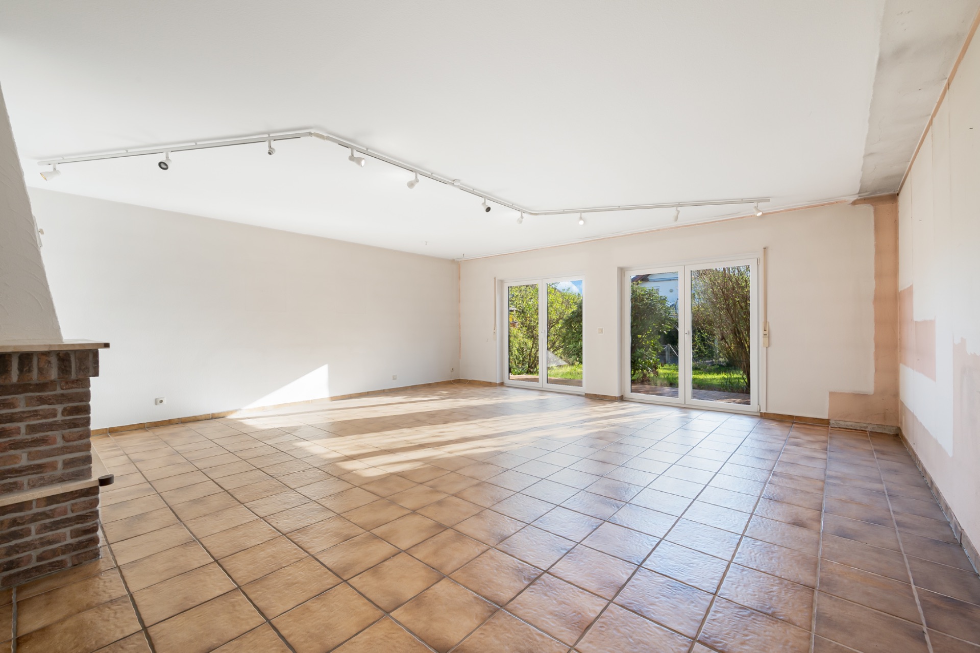 Helles, leeres Wohnzimmer mit Fliesenboden, Kamin und großen Fenstern zum Garten.