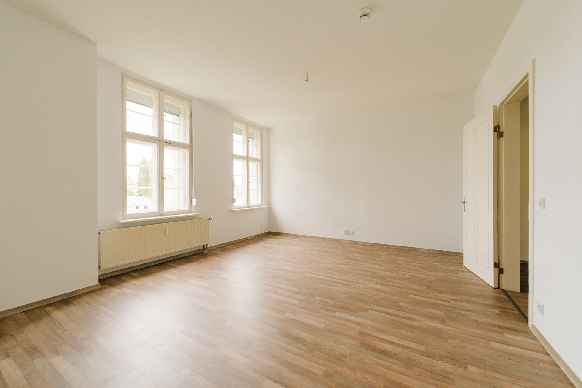 Helles, leeres Zimmer mit drei großen Fenstern, Holzfußboden und weiß gestrichenen Wänden.