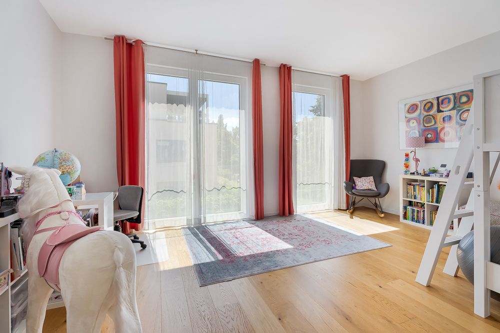 Helles Kinderzimmer mit großen Fenstern, Holzboden, Teppich, Bücherregal und Spielzeugpferd.