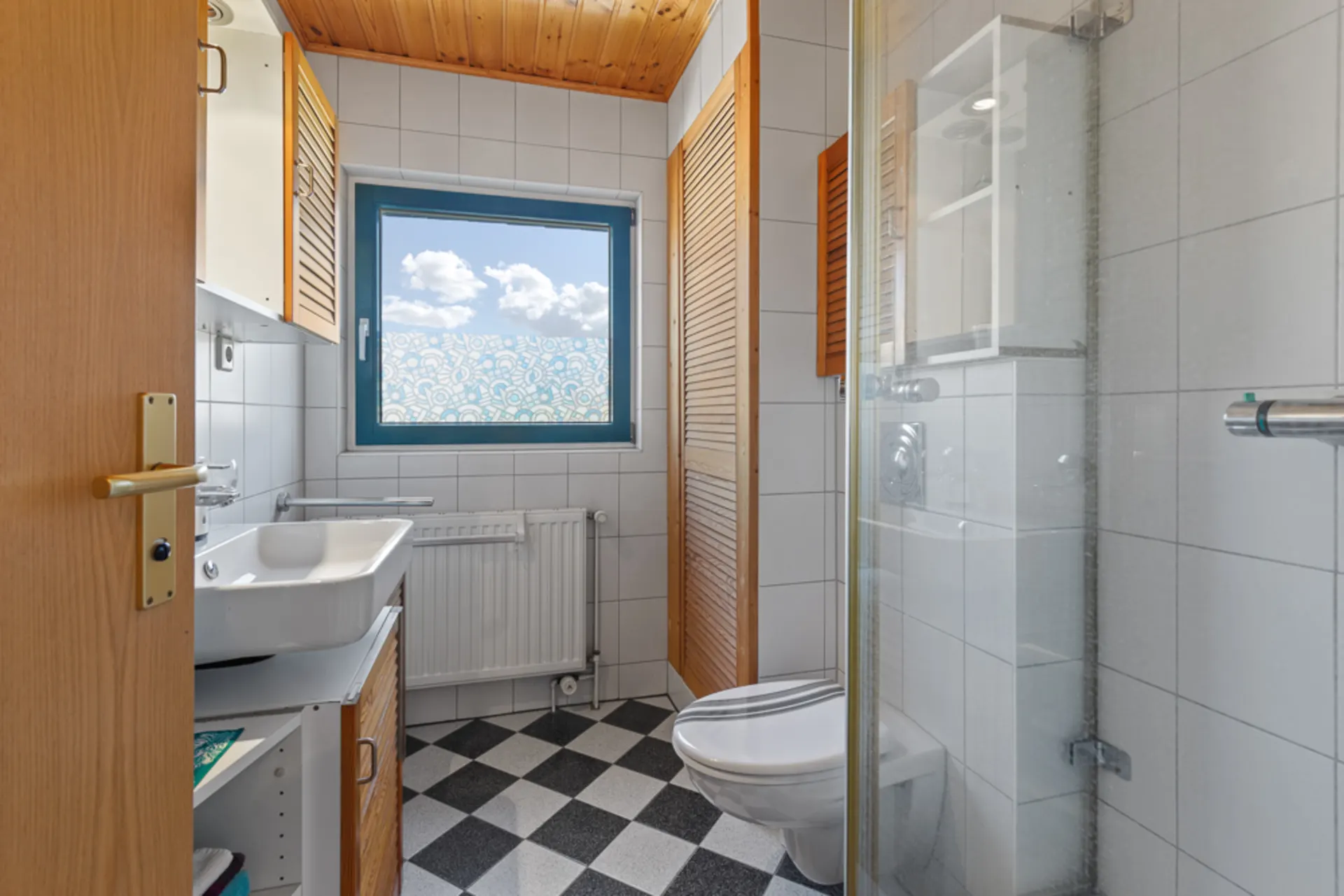 Helles Badezimmer mit WC, Waschbecken, Dusche und Fenster, schwarz-weiß kariertem Boden.