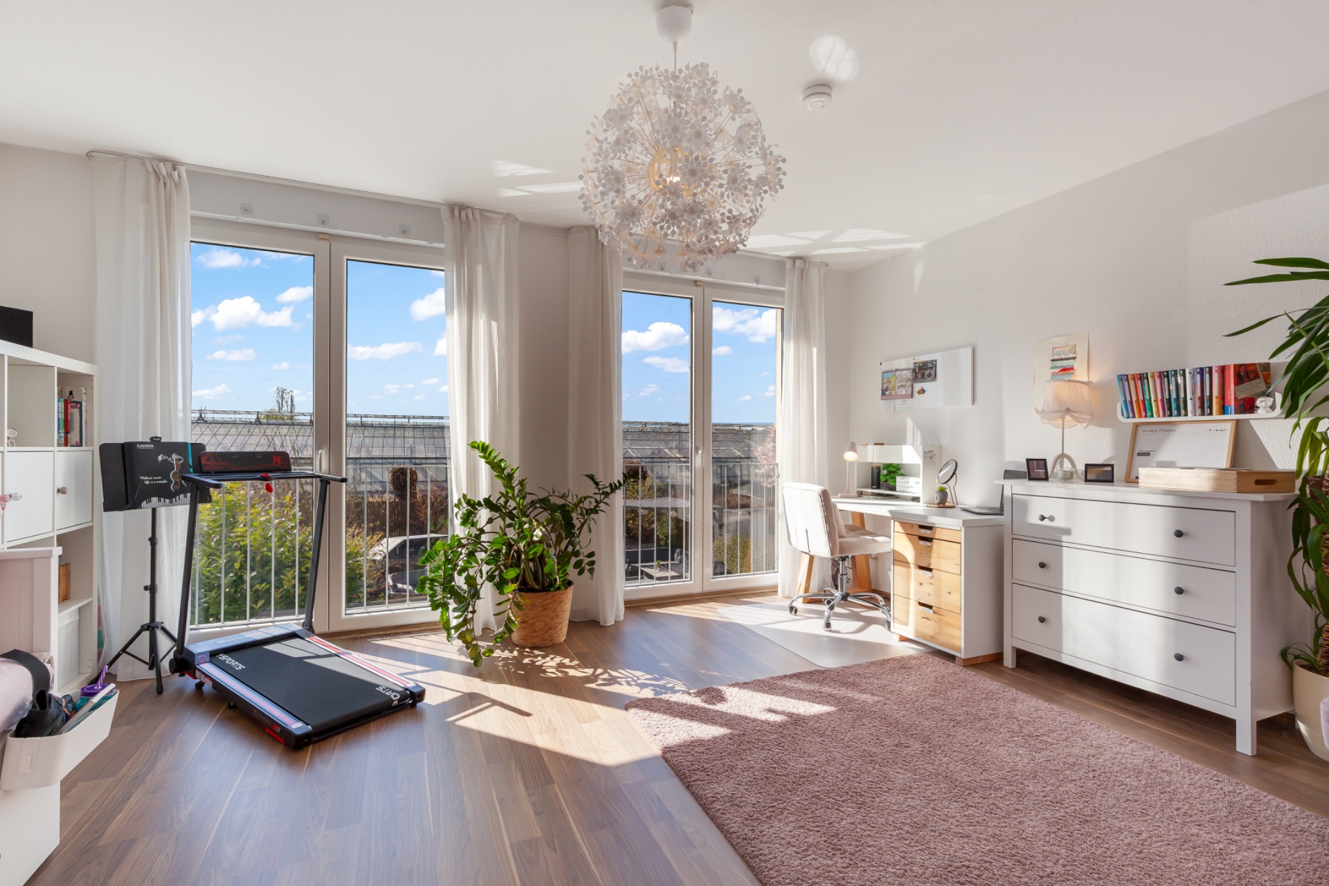 Helles Zimmer mit Schreibtisch, Kommode, Laufband, Regal und großen Fenstern mit Vorhängen.