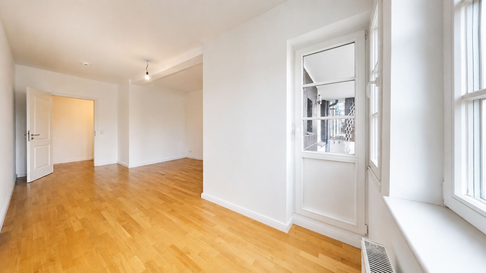 Helles Zimmer mit Holzfußboden, weißen Wänden, Fenster und Glastür nach draußen.
