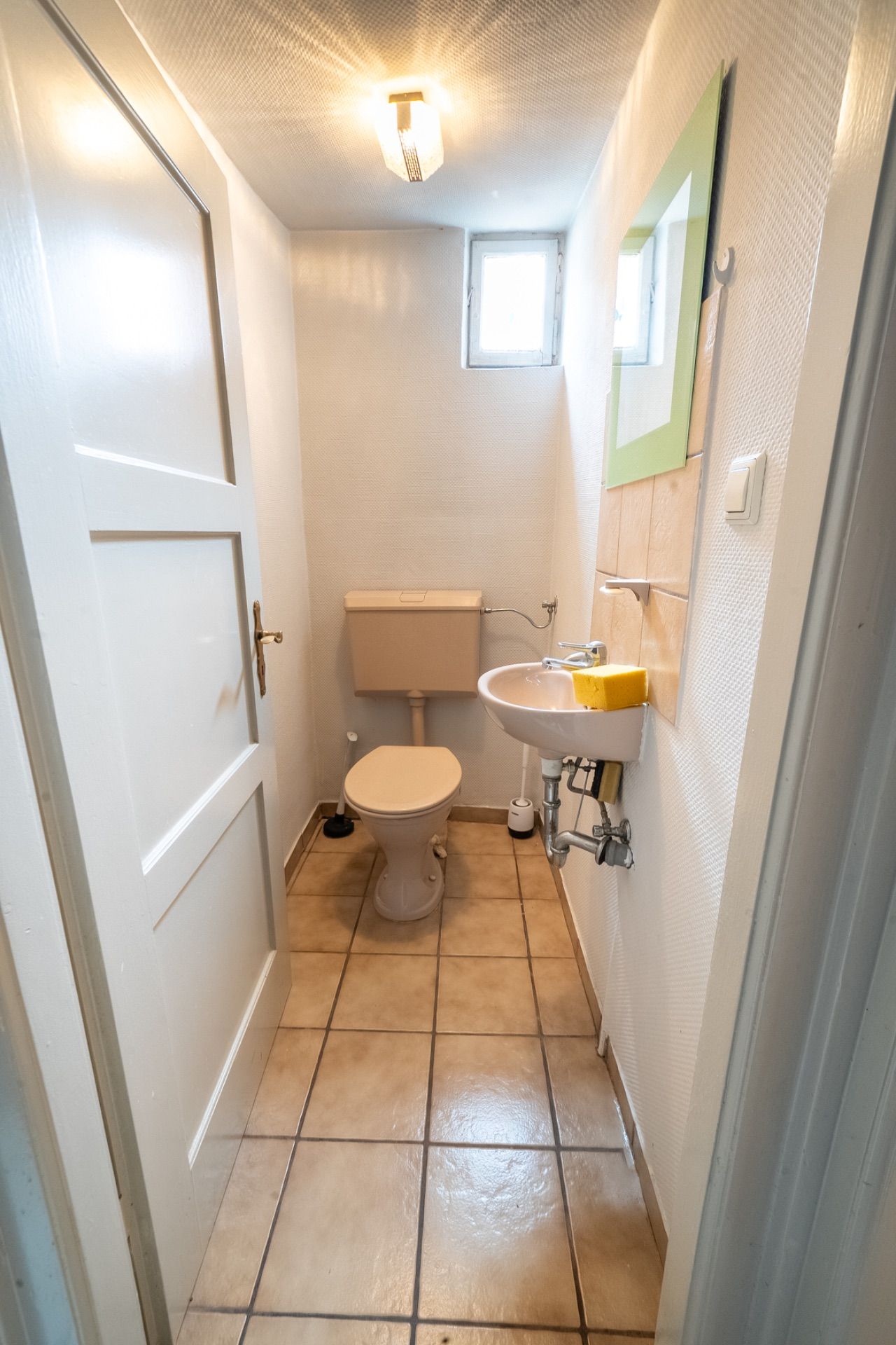 Schmales WC mit Toilette, kleinem Waschbecken, Spiegel und Fenster, gefliestem Boden.