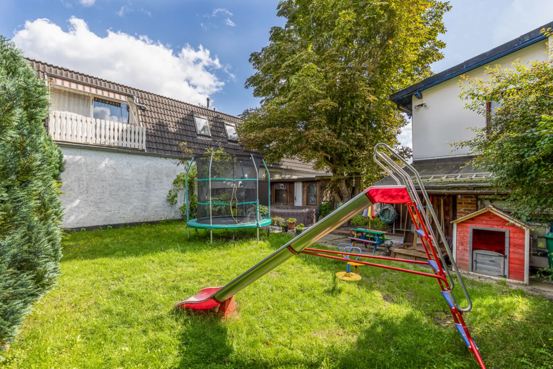Grüner Garten mit Rutsche, Trampolin, Baum und kleinem Spielhaus neben Wohnhäusern.