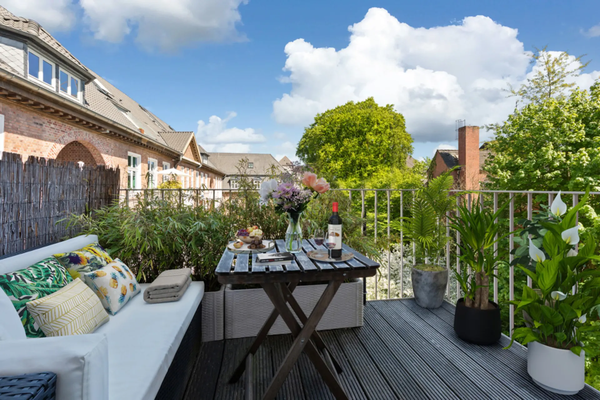 Balkon mit Sofa, Tisch, vielen Pflanzen und Blick auf grüne Bäume und Nachbarhäuser.