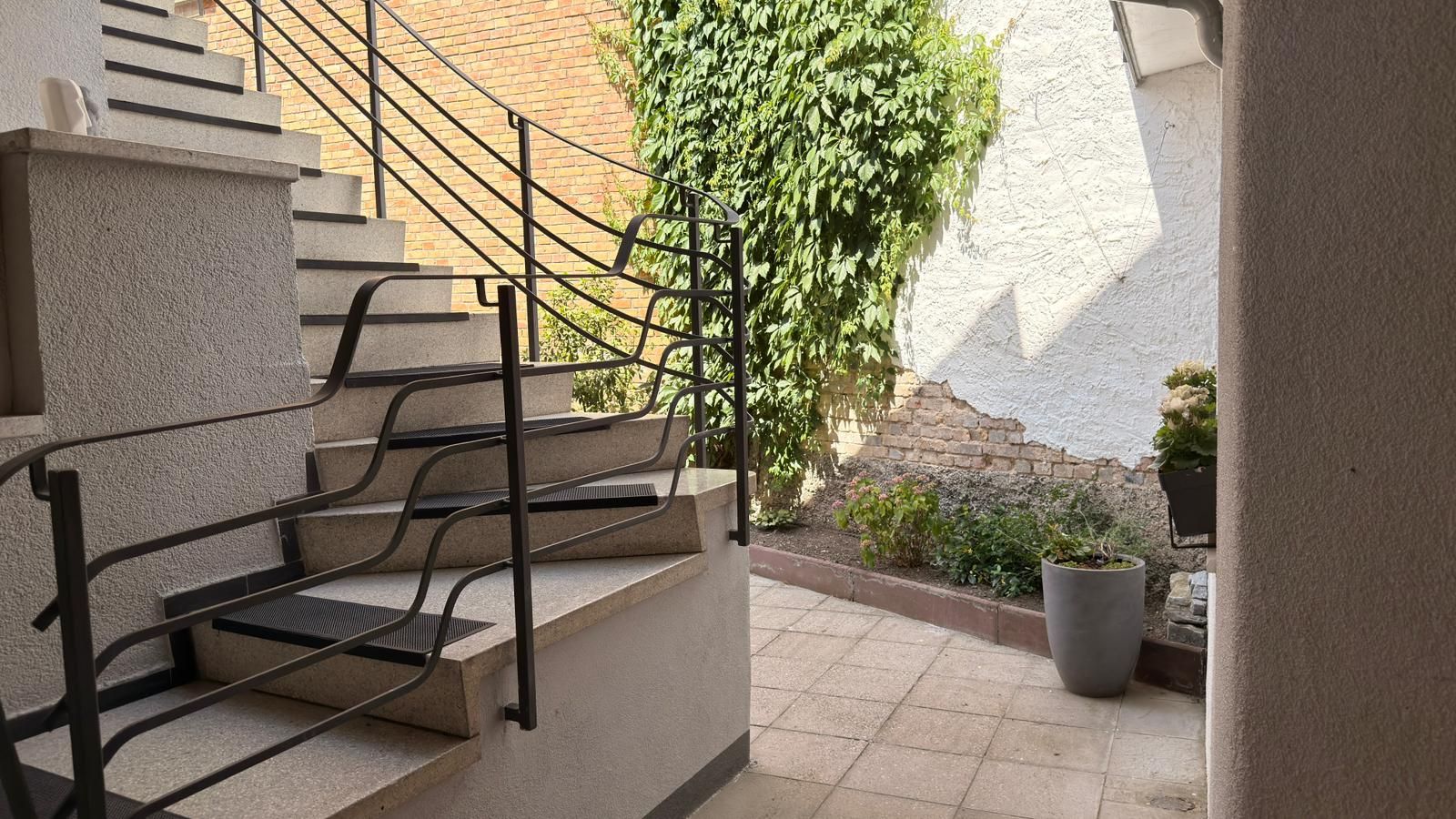 Treppe mit schwarzem Geländer neben kleinem Innenhof mit Pflanzen und Blumentopf.