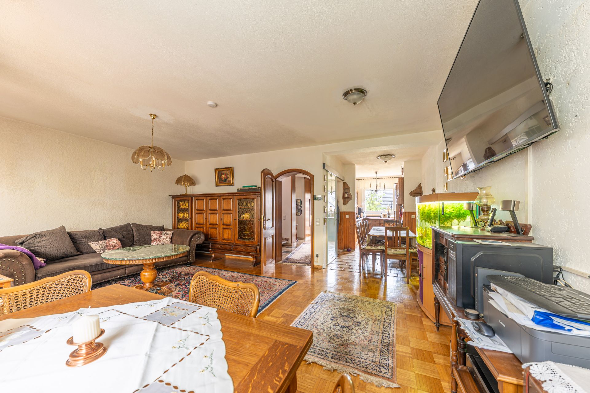 Wohnzimmer mit Esstisch, Sofa, Aquarium, Fernseher und Holzmöbeln, heller Raum.
