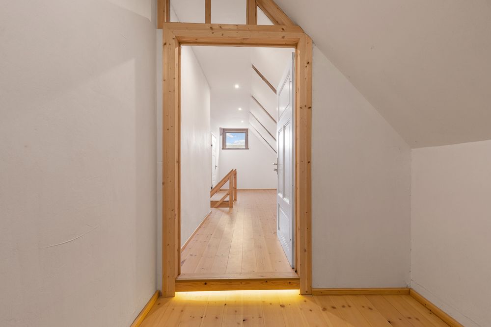 Schmaler Flur mit Holzboden und Dachschräge, am Ende ein Fenster und eine weiße Tür.