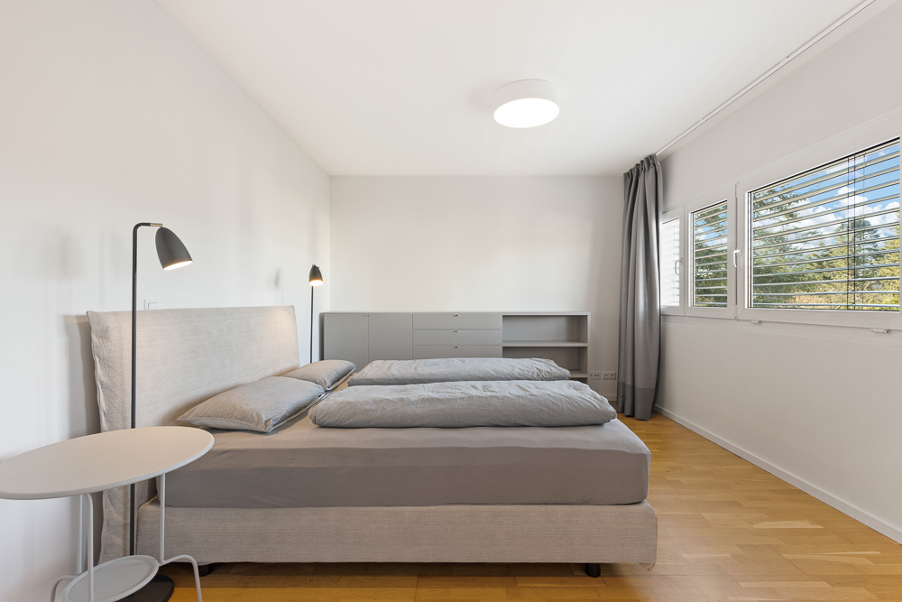 Helles Schlafzimmer mit Doppelbett, Nachttisch, Stehlampen, Kommode und Fenstern mit Jalousien.