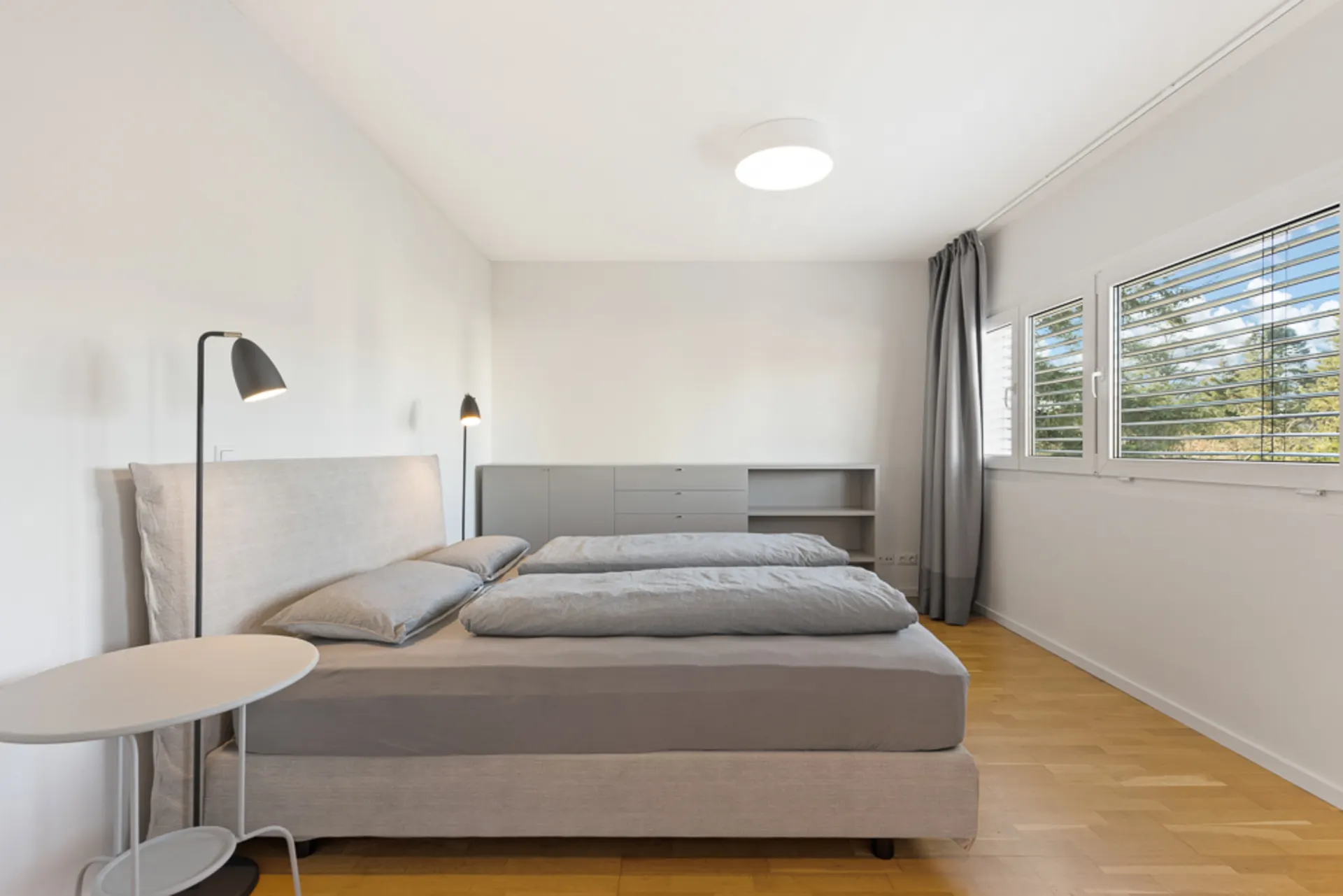 Helles Schlafzimmer mit Doppelbett, Nachttisch, Stehlampen, Kommode und Fenstern mit Jalousien.