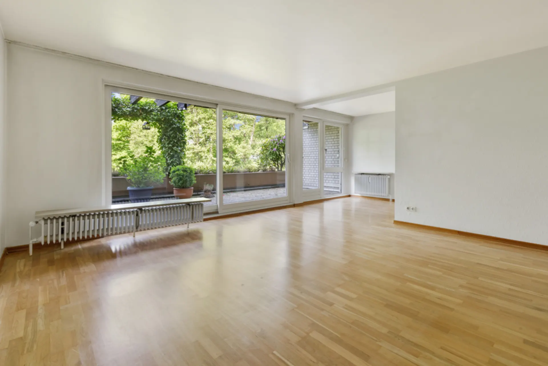 Helles, leeres Wohnzimmer mit Holzfußboden und großen Fenstern zur Terrasse.