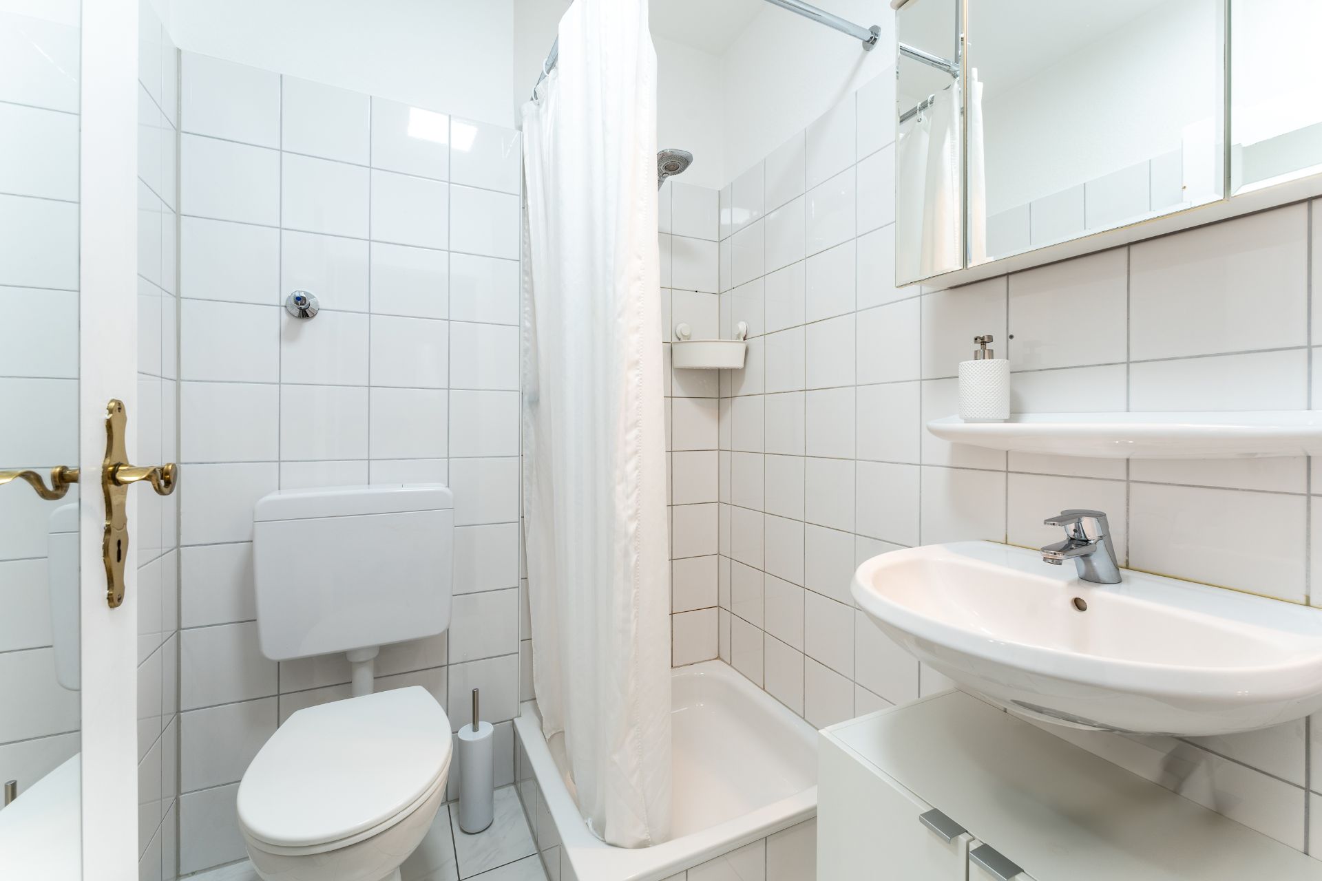 Kleines Badezimmer mit Dusche, Toilette und Waschbecken, alles mit weißen Fliesen.