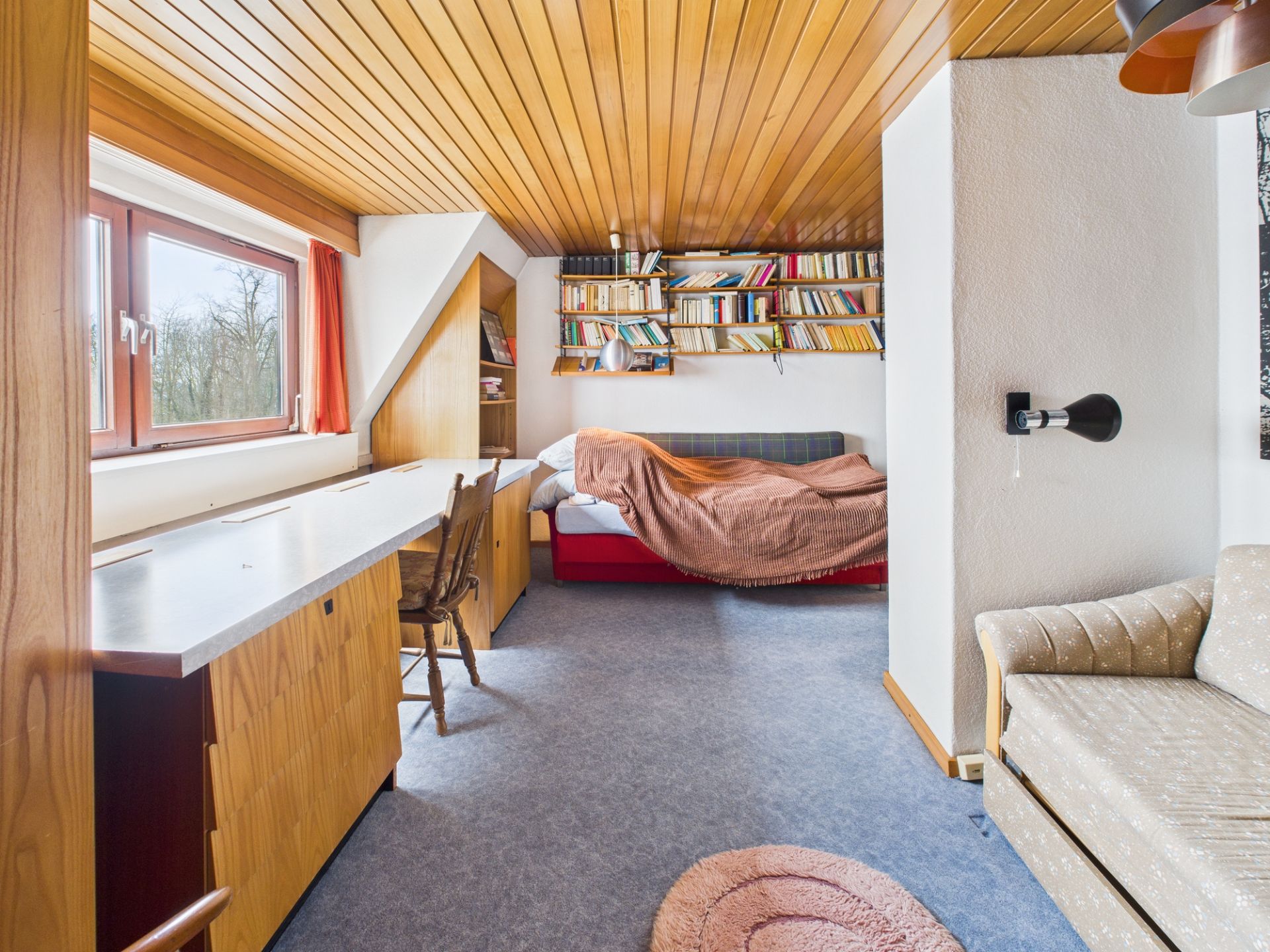 Helles Zimmer mit Holzmöbeln, Schreibtisch, Bett, Sofa und Bücherregal an der Wand.