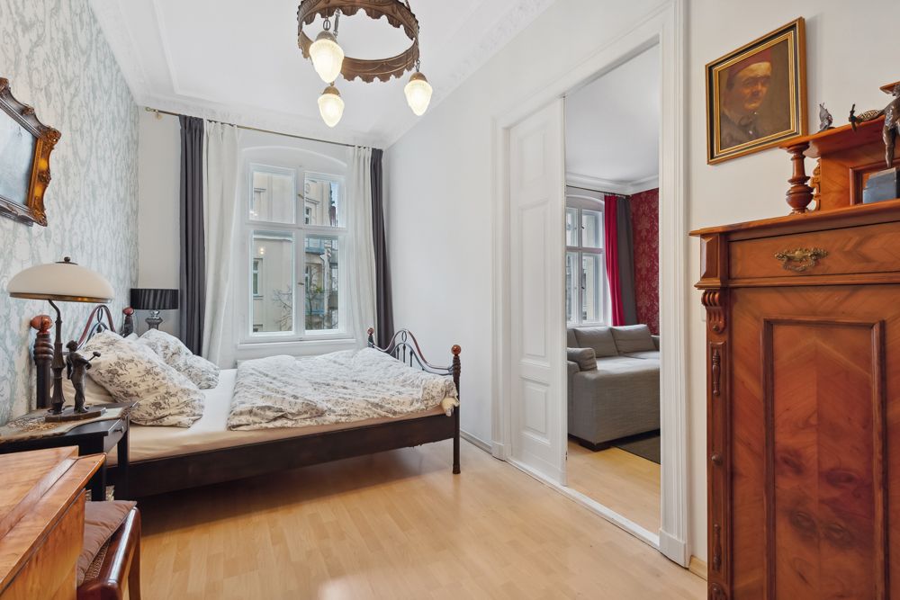 Helles Schlafzimmer mit Doppelbett, Holzschrank und Blick in ein Wohnzimmer.