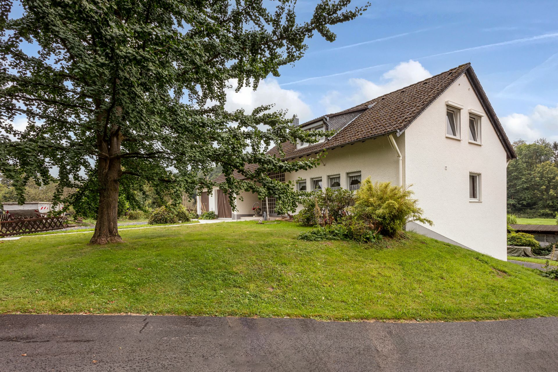Weißes Einfamilienhaus mit braunem Dach, großer Baum und Rasen im Vordergrund.