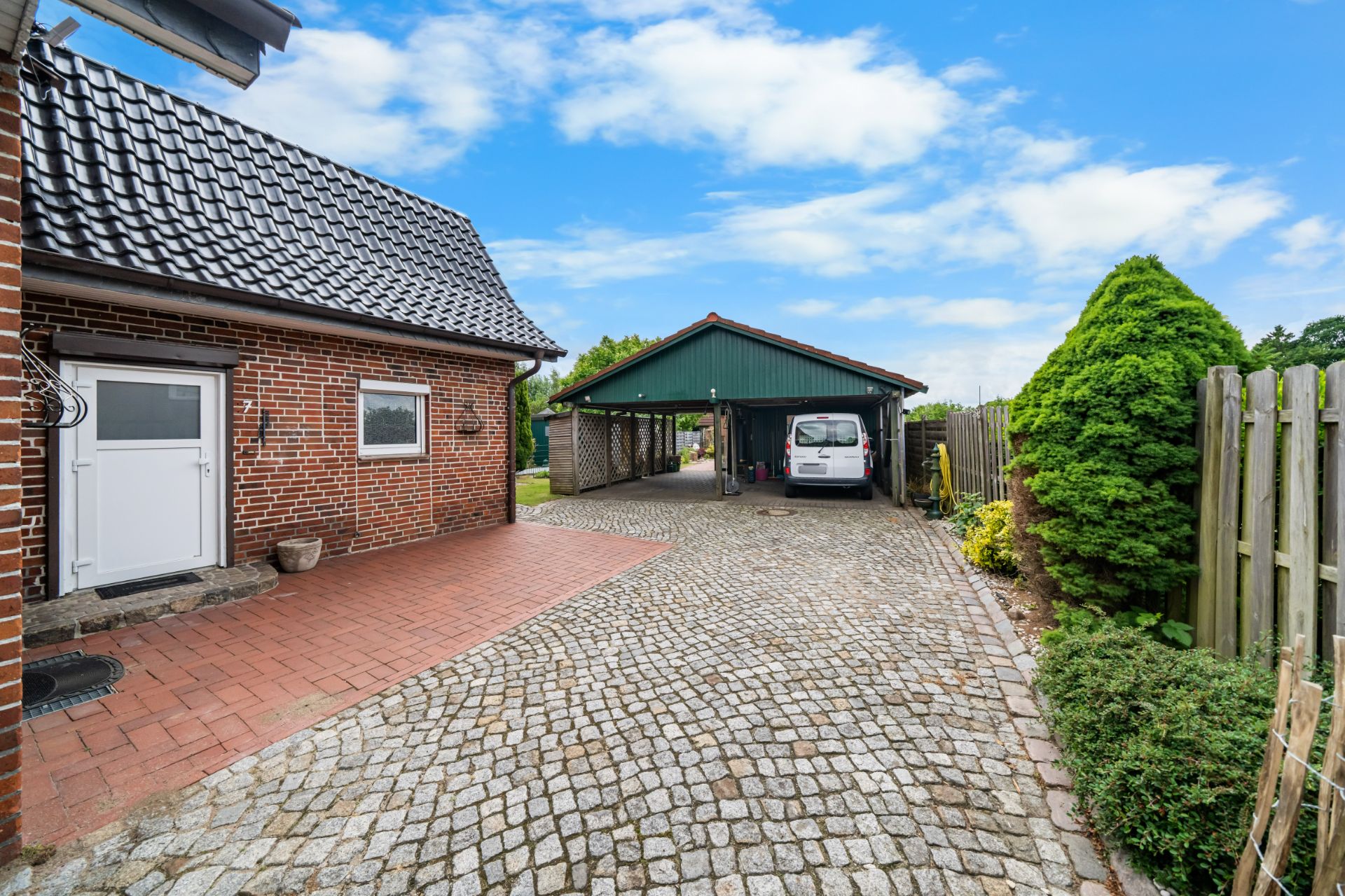 Gepflasterter Hof mit Haus, Garage, Auto und grünen Pflanzen an einem sonnigen Tag.