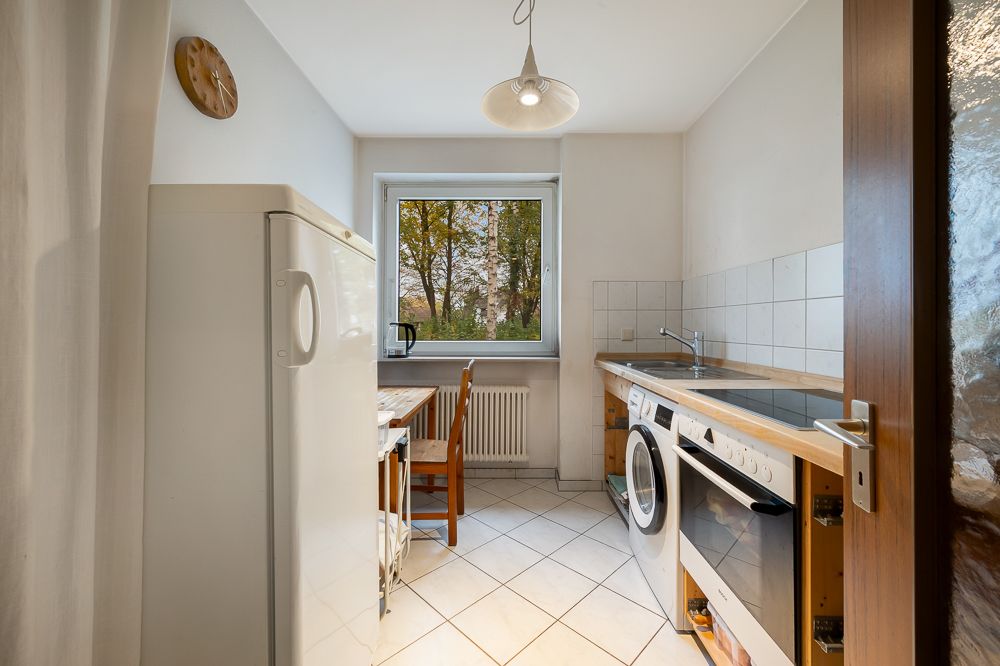 Helle Küche mit Kühlschrank, Esstisch, Waschmaschine und großem Fenster mit Blick ins Grüne.