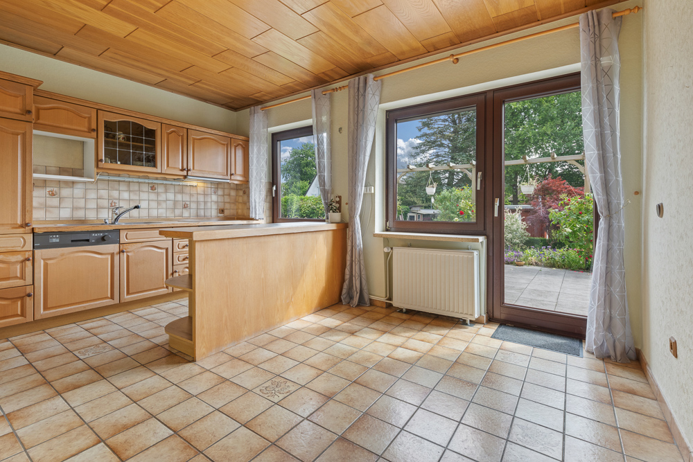 Helle Küche mit Holzmöbeln, Fliesenboden und Zugang zur Terrasse mit Gartenblick.