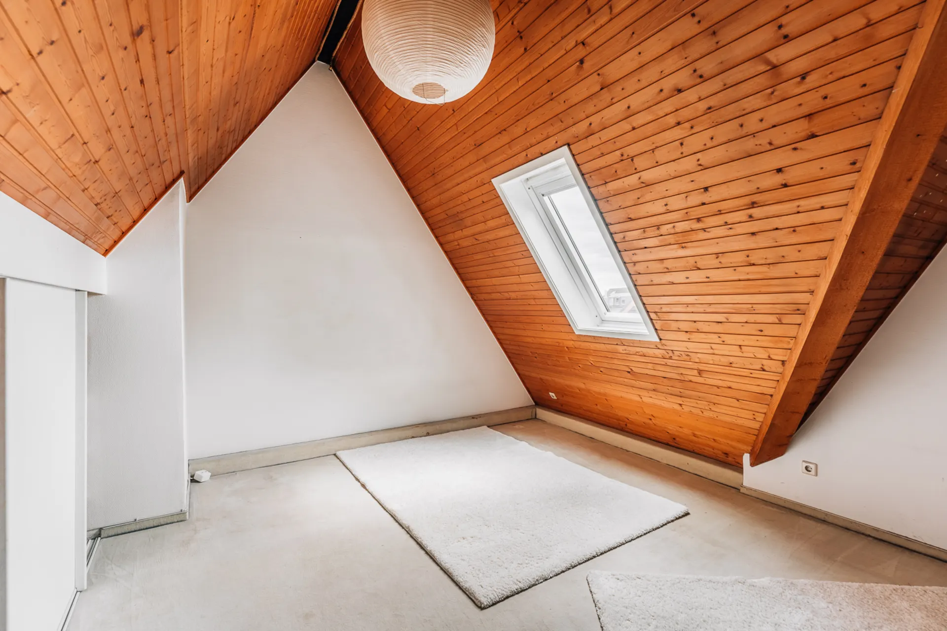 Dachzimmer mit Holzdecke, Dachfenster und zwei hellen Teppichen auf dem Boden.