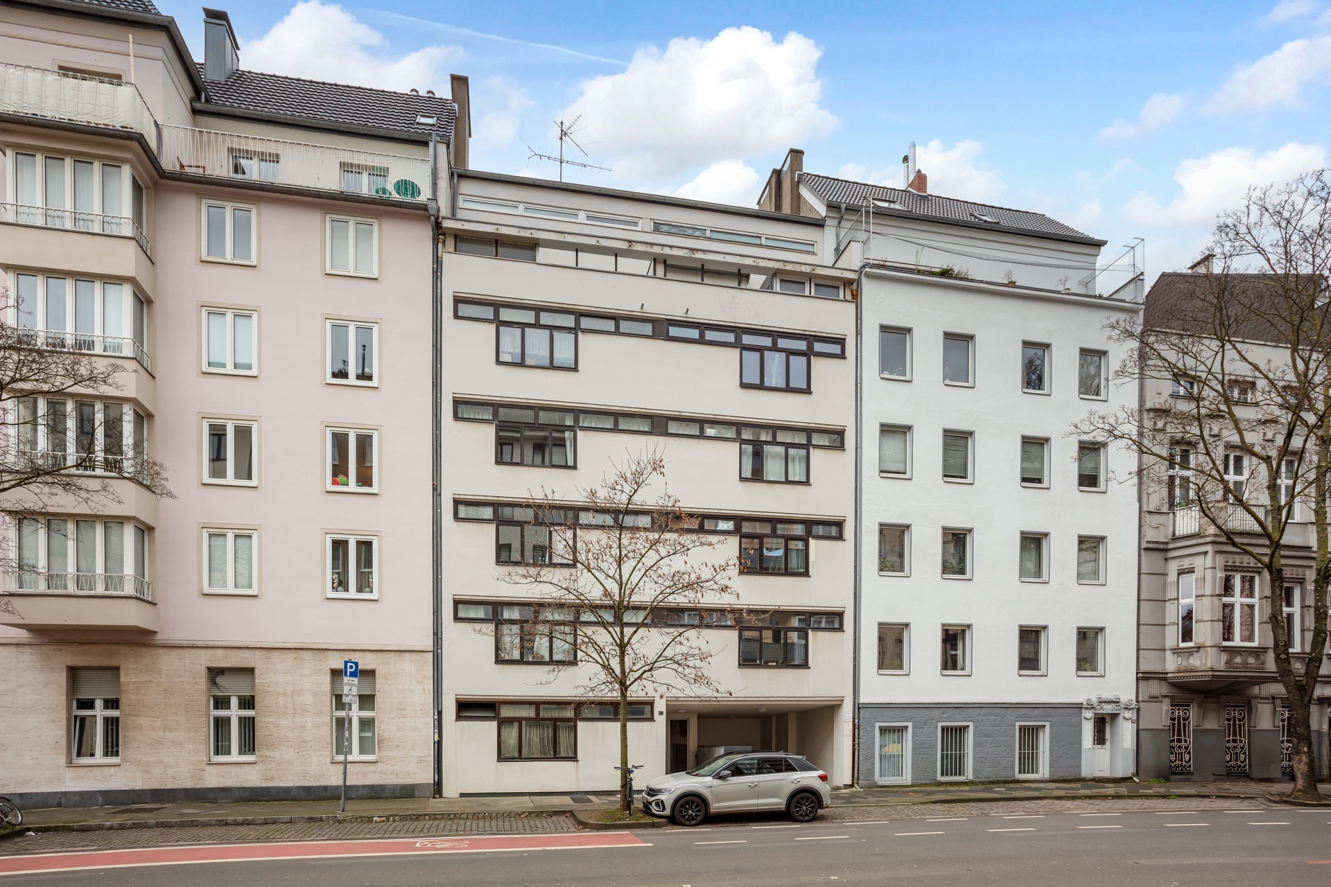 Mehrfamilienhaus mit mehreren Etagen und großen Fenstern an einer Straße, ein Auto parkt davor.