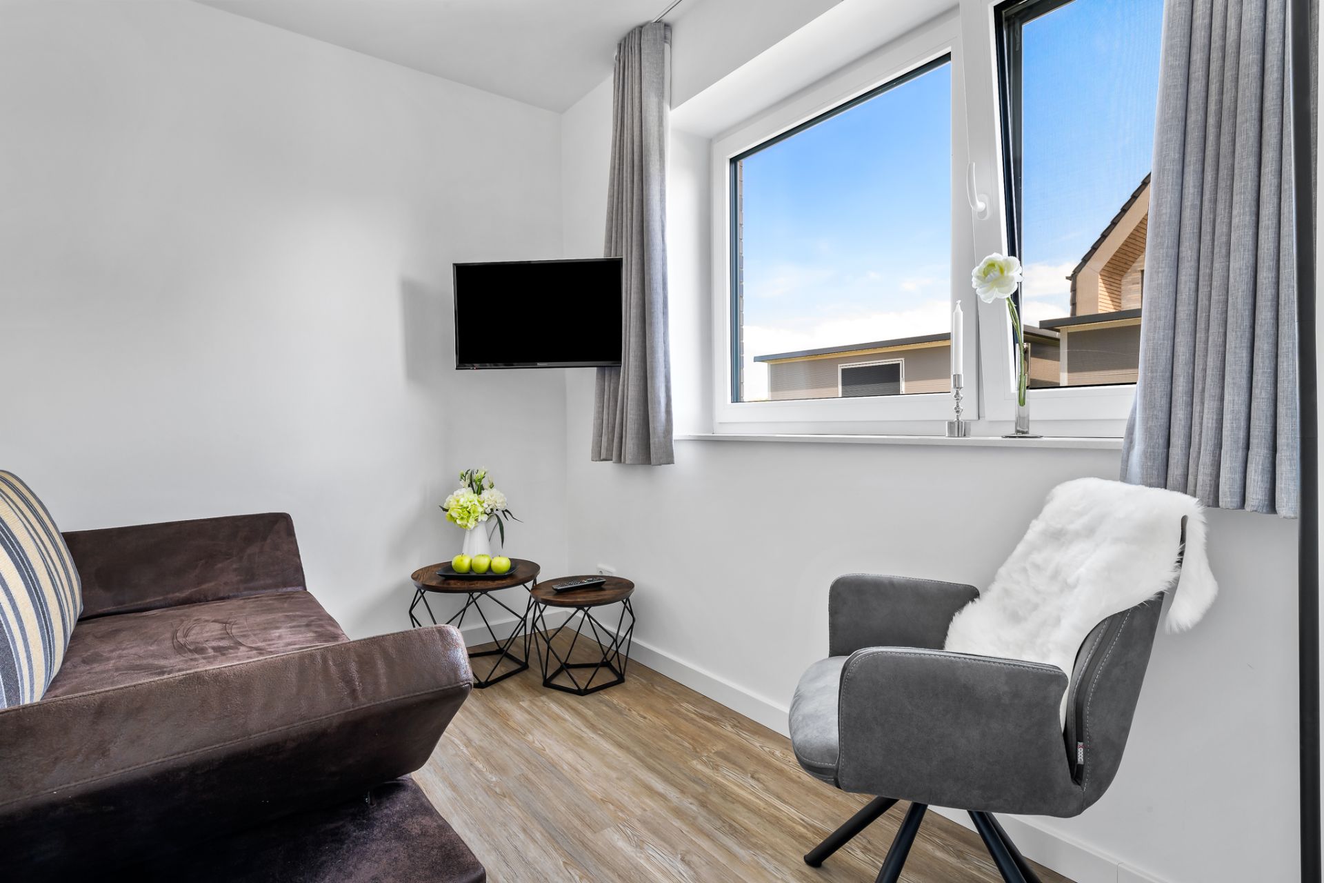 Kleines Wohnzimmer mit Sofa, Sessel, Fernseher, zwei Tischen und großem Fenster.