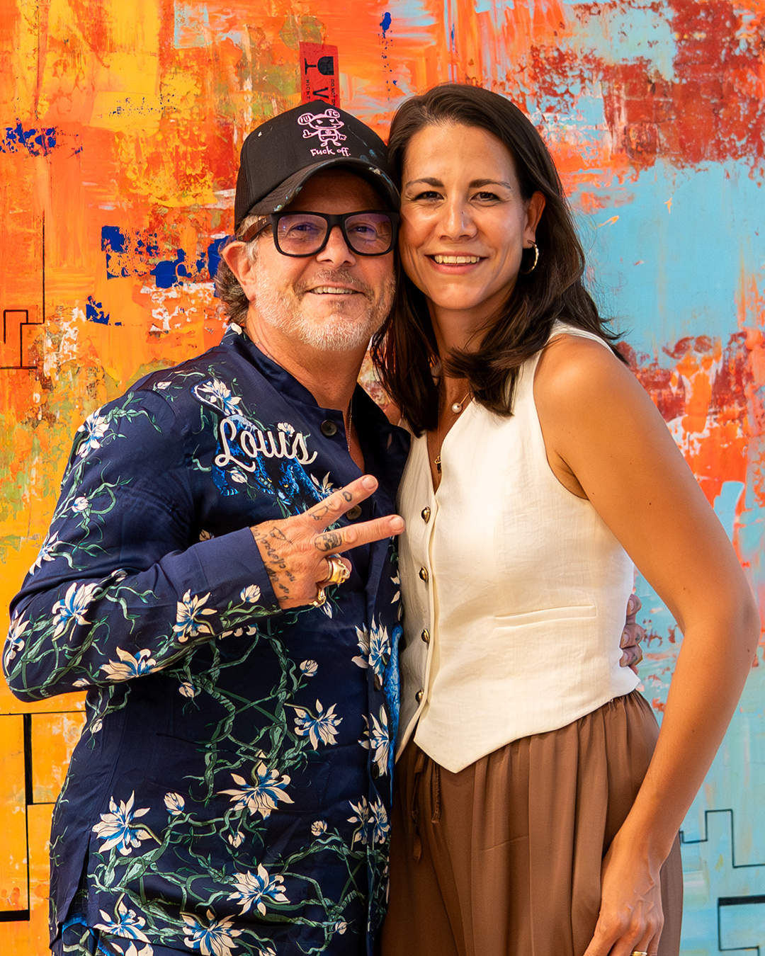 Zwei lächelnde Personen stehen vor einer bunten, abstrakten Wand.