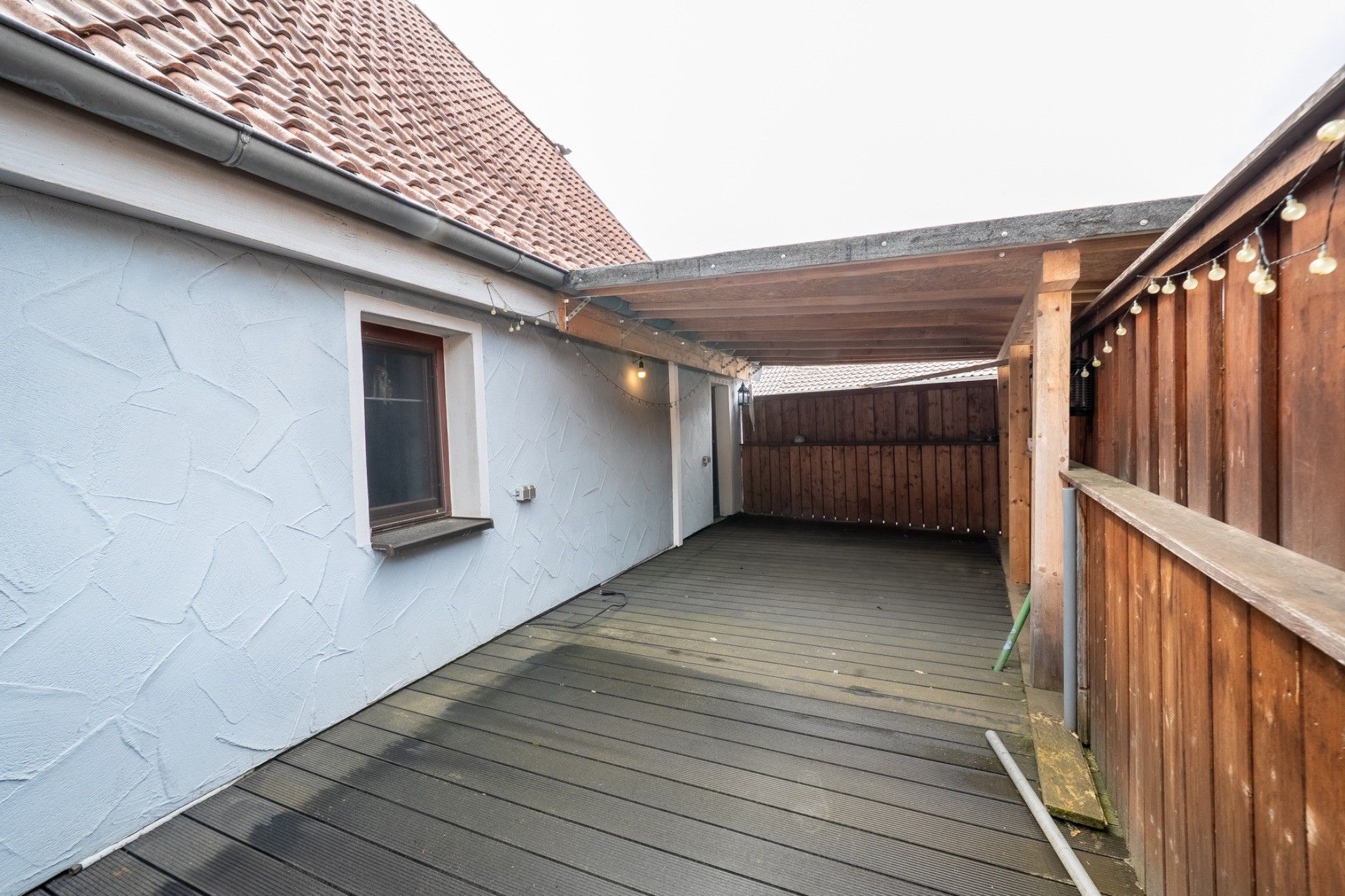 Überdachte Terrasse mit Holzboden, Holzzaun und Lichterkette an einem Wohnhaus.