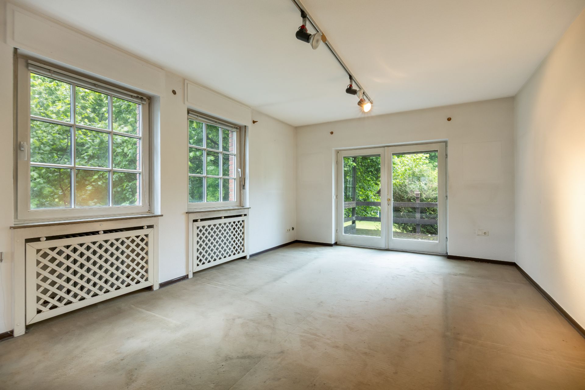 Leeres Zimmer mit zwei Fenstern und Glastür zum Garten, helle Wände und Deckenleuchte.