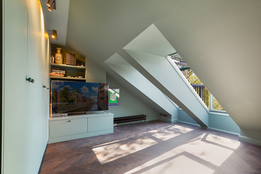 Helles Dachzimmer mit schrägen Fenstern, Fernseher, Regalen und Holzfußboden.