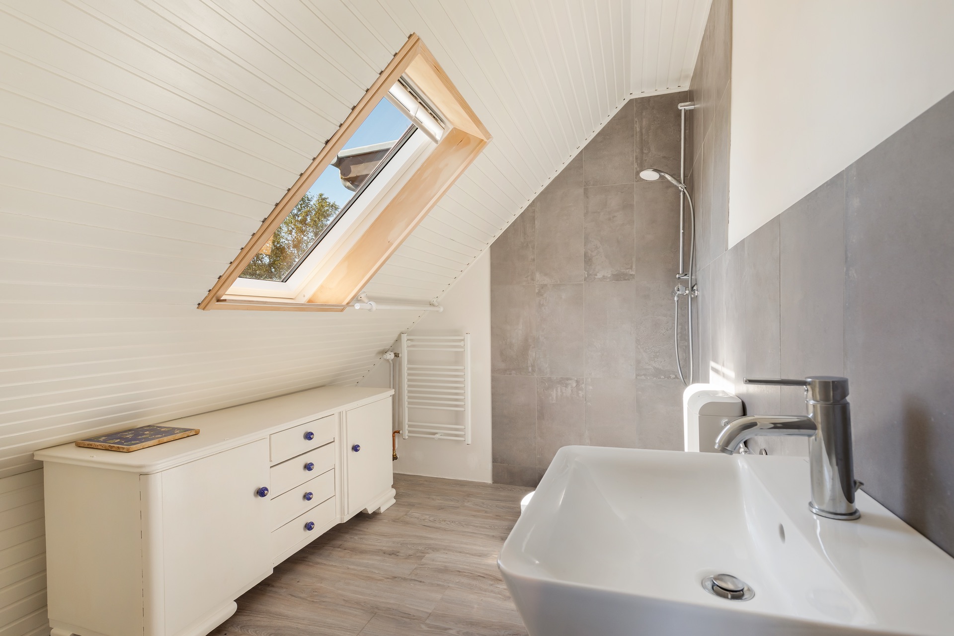 Helles Badezimmer mit Dachfenster, Waschbecken, Dusche und weißer Kommode.