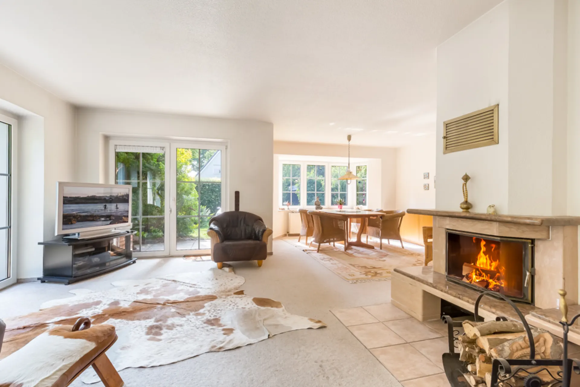 Helles Wohnzimmer mit Kamin, Fernseher, Lederstuhl, Teppich und Essbereich mit großem Fenster.