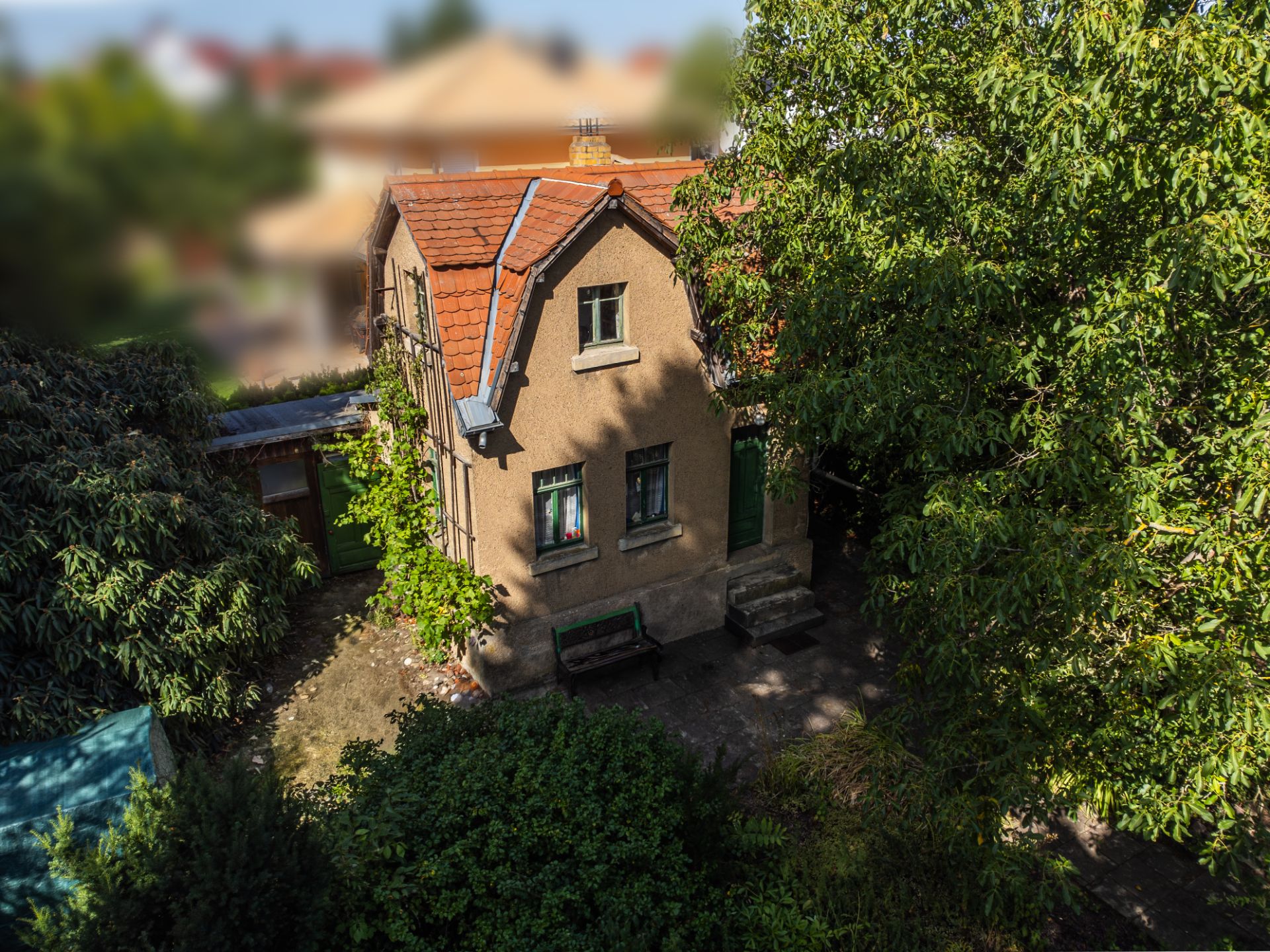Kleines Haus mit rotem Dach, Fensterläden und Bank im schattigen Garten, umgeben von Bäumen.