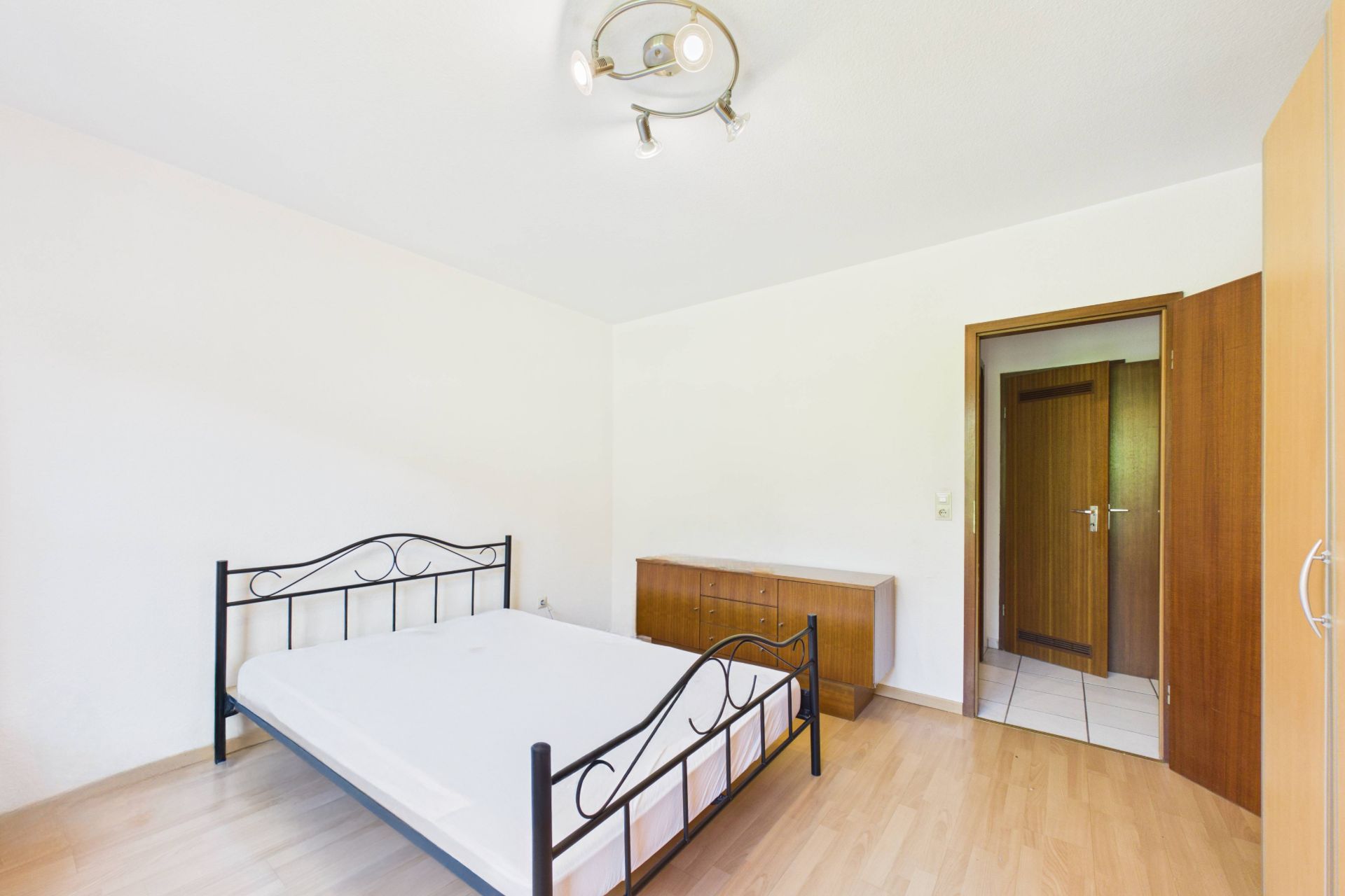 Helles Schlafzimmer mit Metallbett, Kommode und Kleiderschrank auf Laminatboden.
