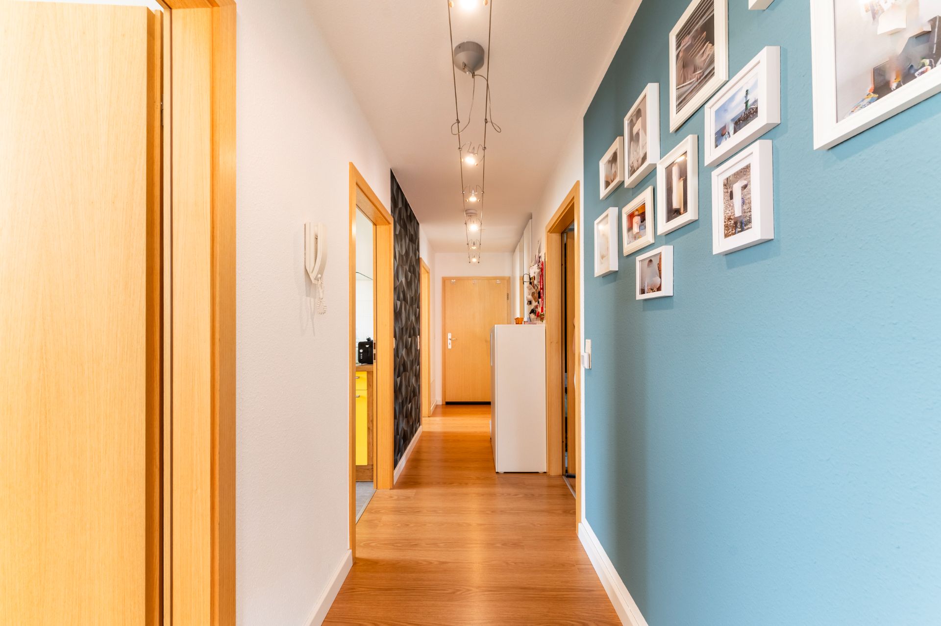 Heller Flur mit Holzboden, blauer Wand und mehreren gerahmten Fotos an der Wand.