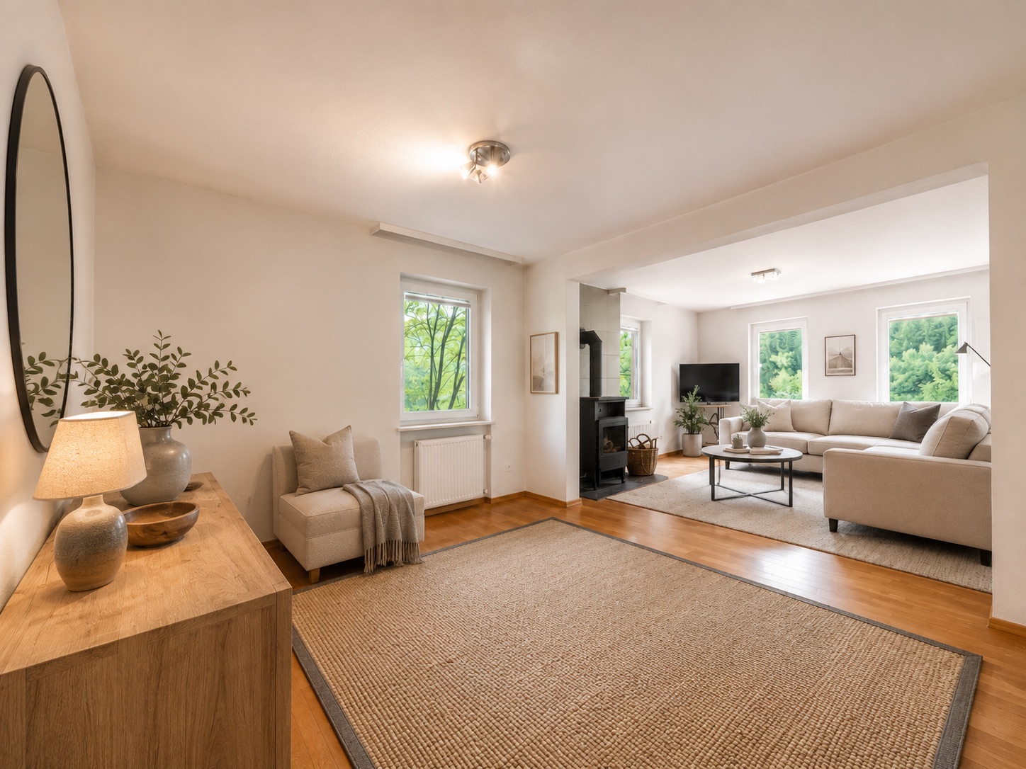 Helles Wohnzimmer mit Sofa, Sessel, Teppich und Holzmöbeln, große Fenster bieten Blick ins Grüne.