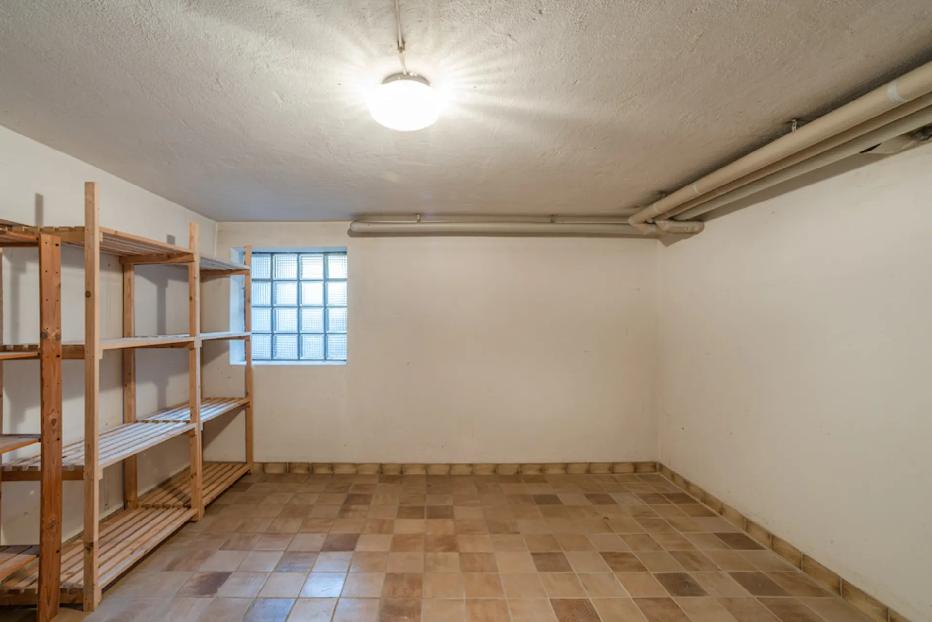 Kellerraum mit Fliesenboden, Regal an der Wand und kleinem Fenster, helle Deckenlampe.