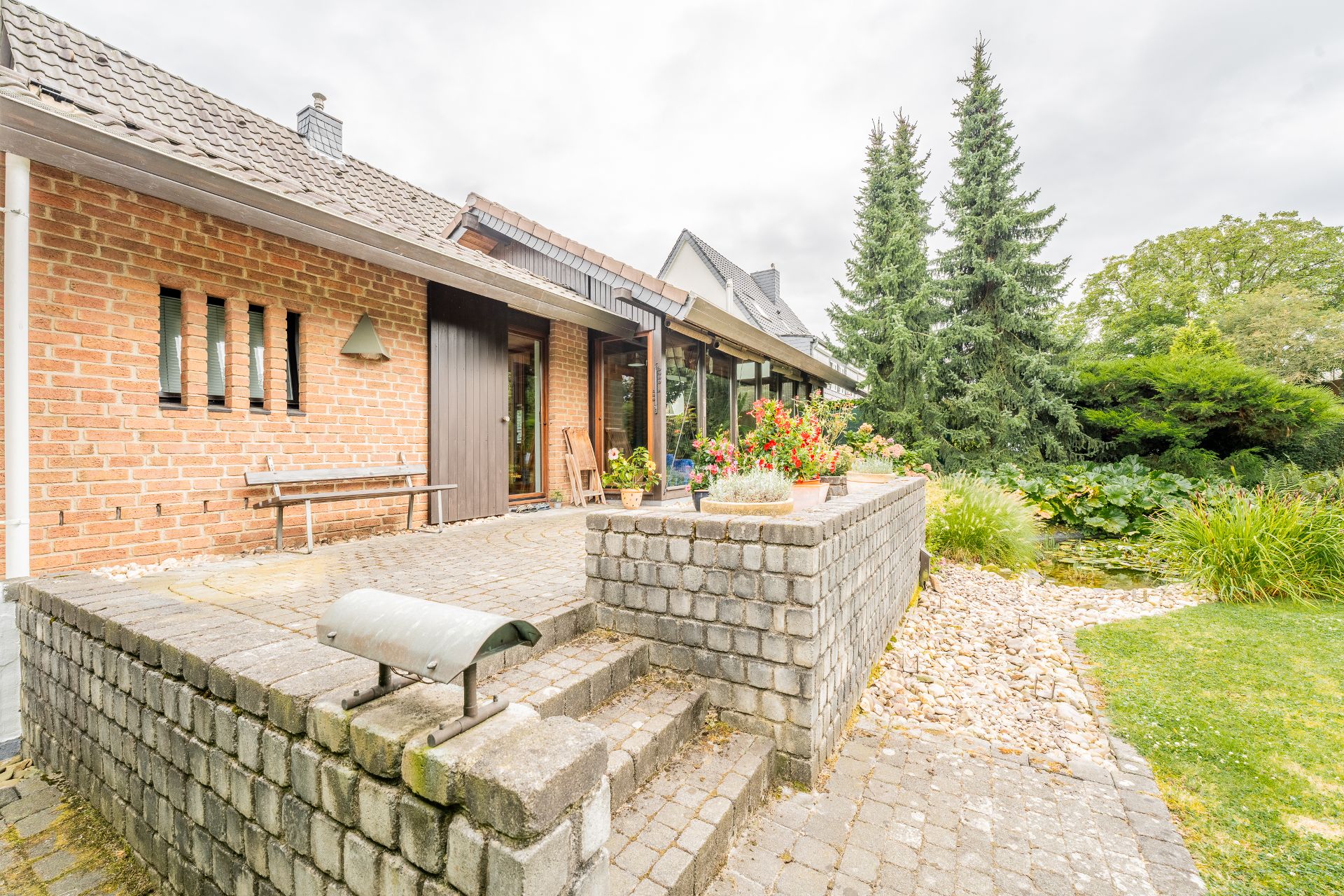 Gepflegte Terrasse aus Stein mit Blumenkübeln und Garten, im Hintergrund hohe Tannen.
