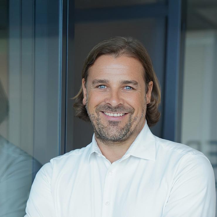 Mann mit Bart und weißem Hemd steht lächelnd mit verschränkten Armen vor Glaswand.