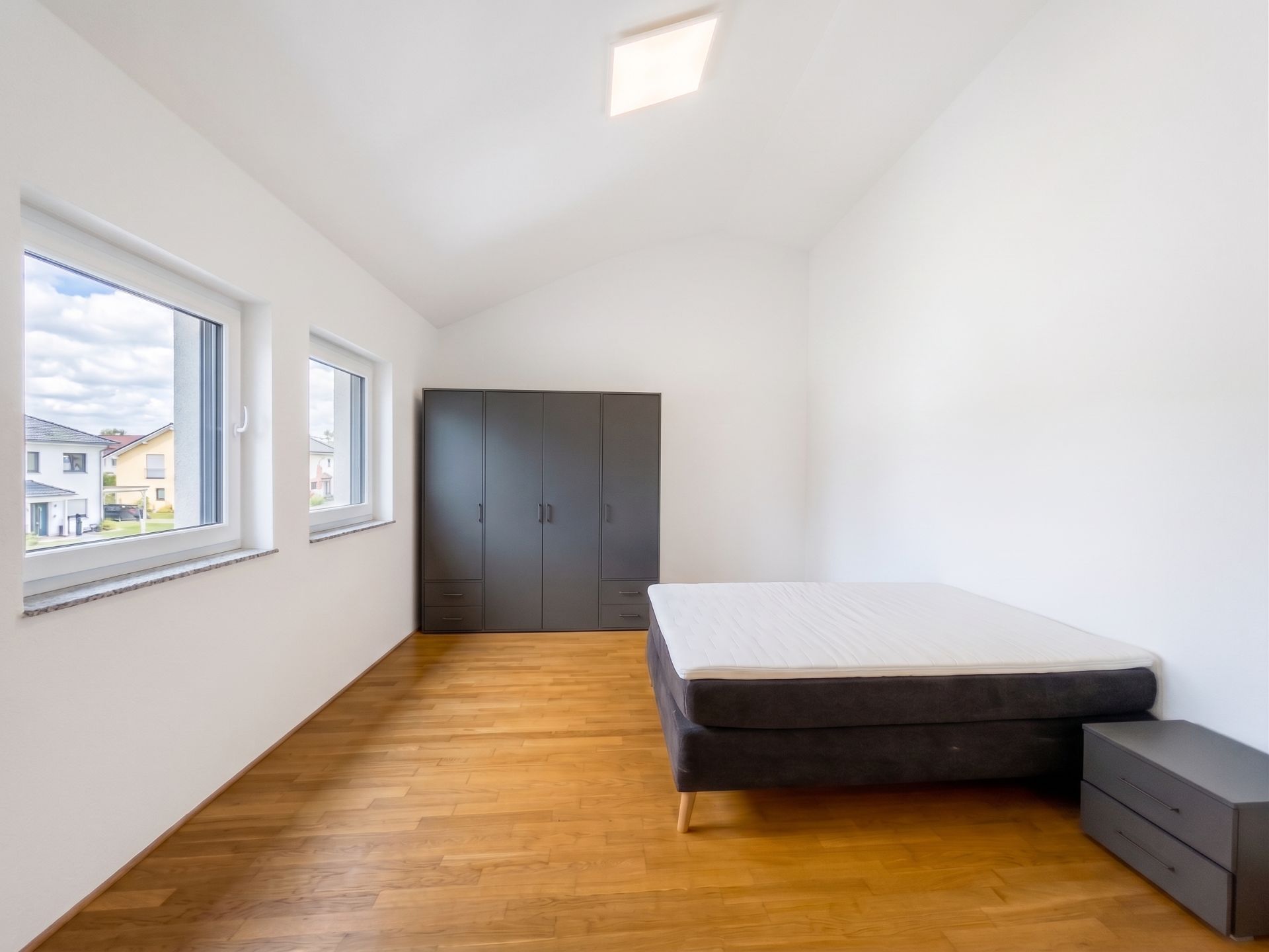 Helles Schlafzimmer mit drei Fenstern, einem Bett, Nachttisch und großem Kleiderschrank auf Holzfußboden.