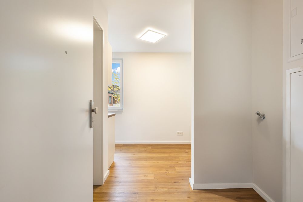 Heller Flur mit Holzboden, weißer Wand und Fenster im Hintergrund.