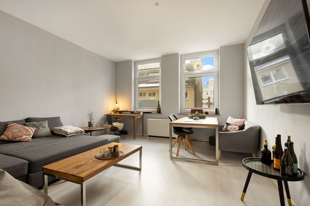 Wohnzimmer mit grauem Sofa, Holztisch, Esstisch, Schreibtisch und großem Fenster.