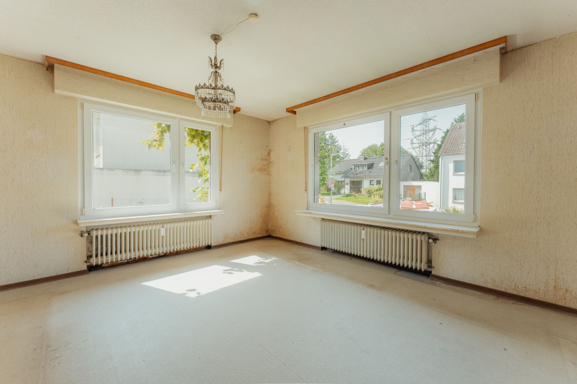 Heller, leerer Raum mit zwei großen Fenstern, Heizkörpern und Kronleuchter.