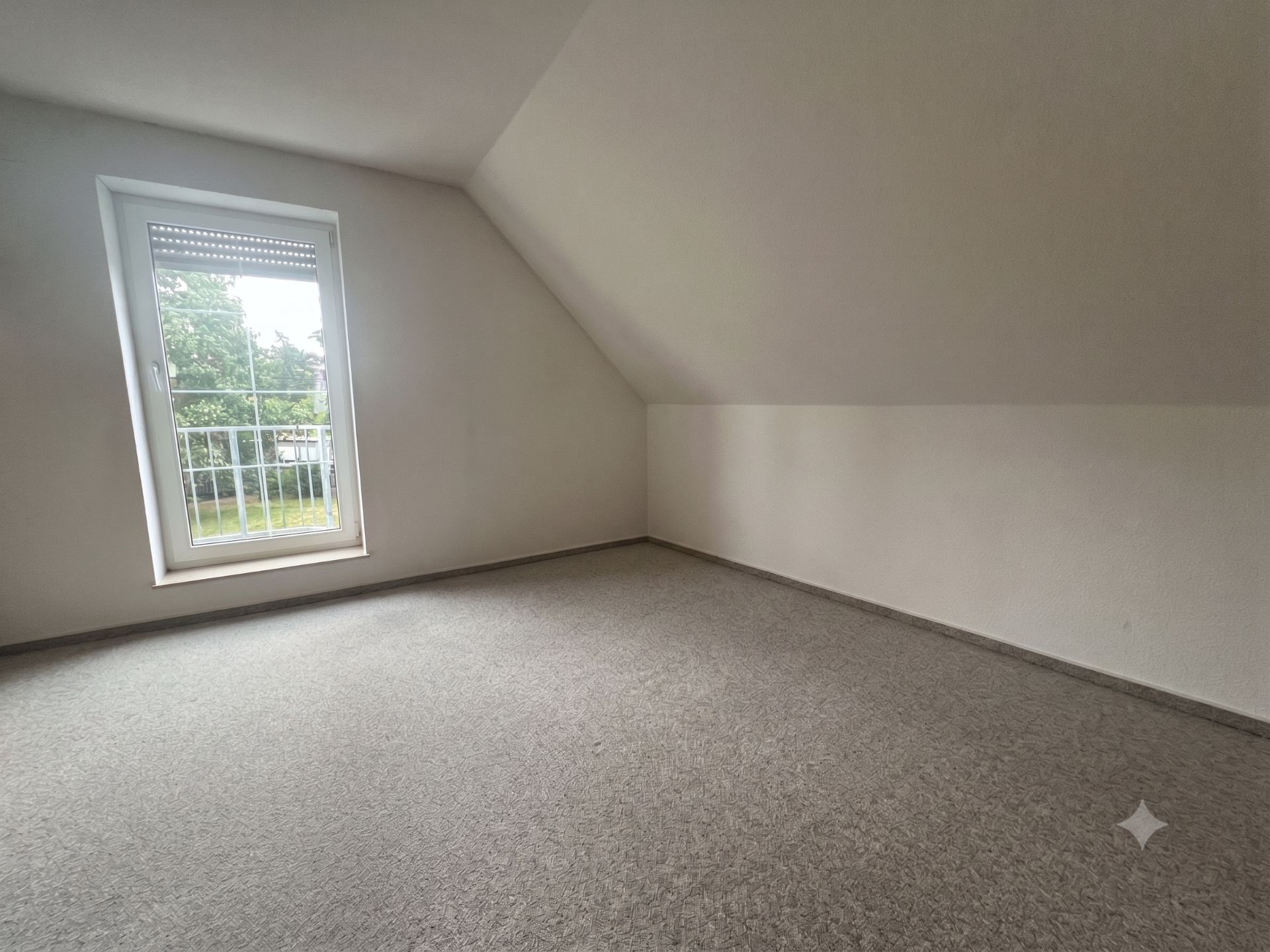 Leerer Raum mit Teppichboden und großem Fenster unter einer Dachschräge.