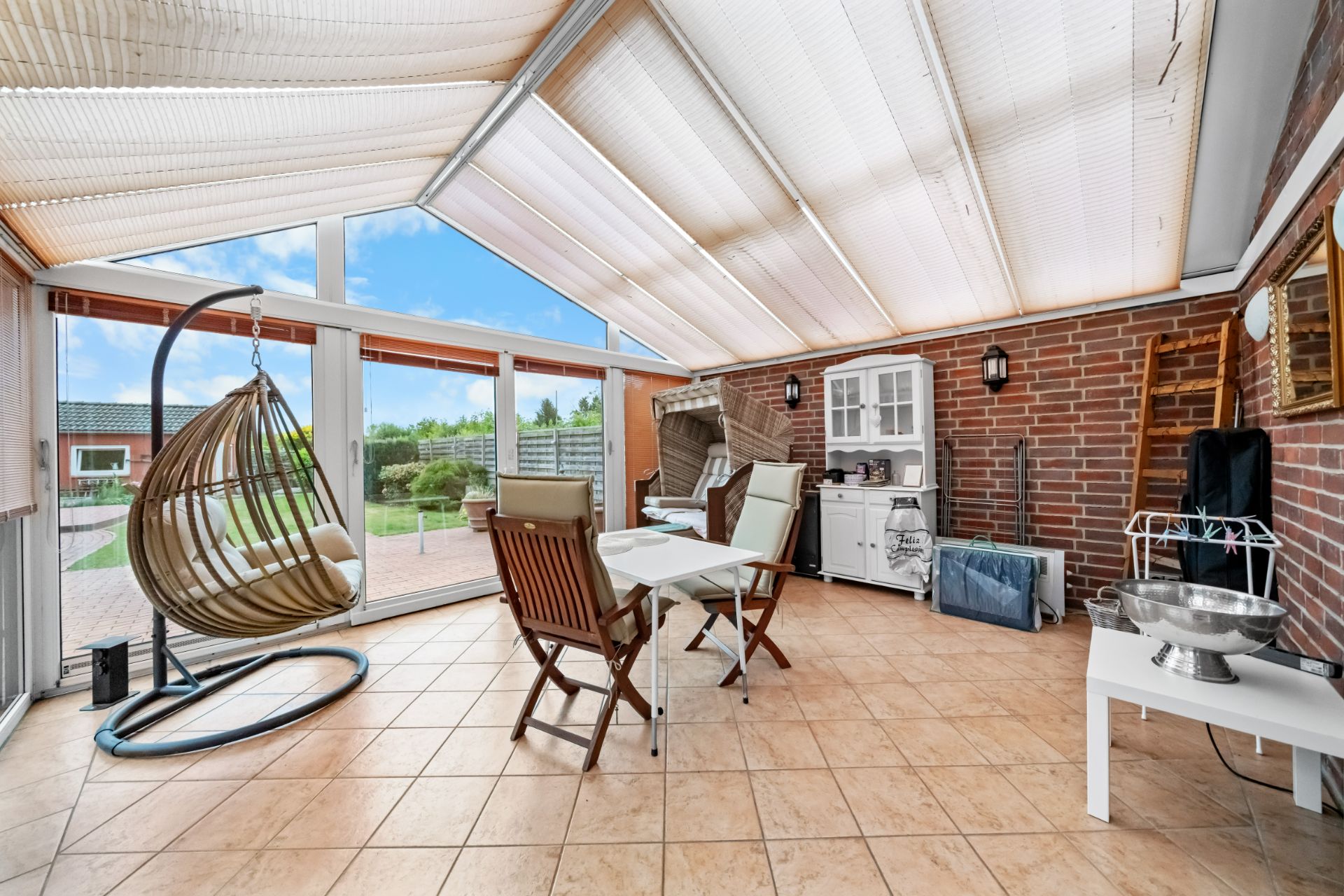 Heller Wintergarten mit Esstisch, Hängesessel und Sideboard auf gefliestem Boden.