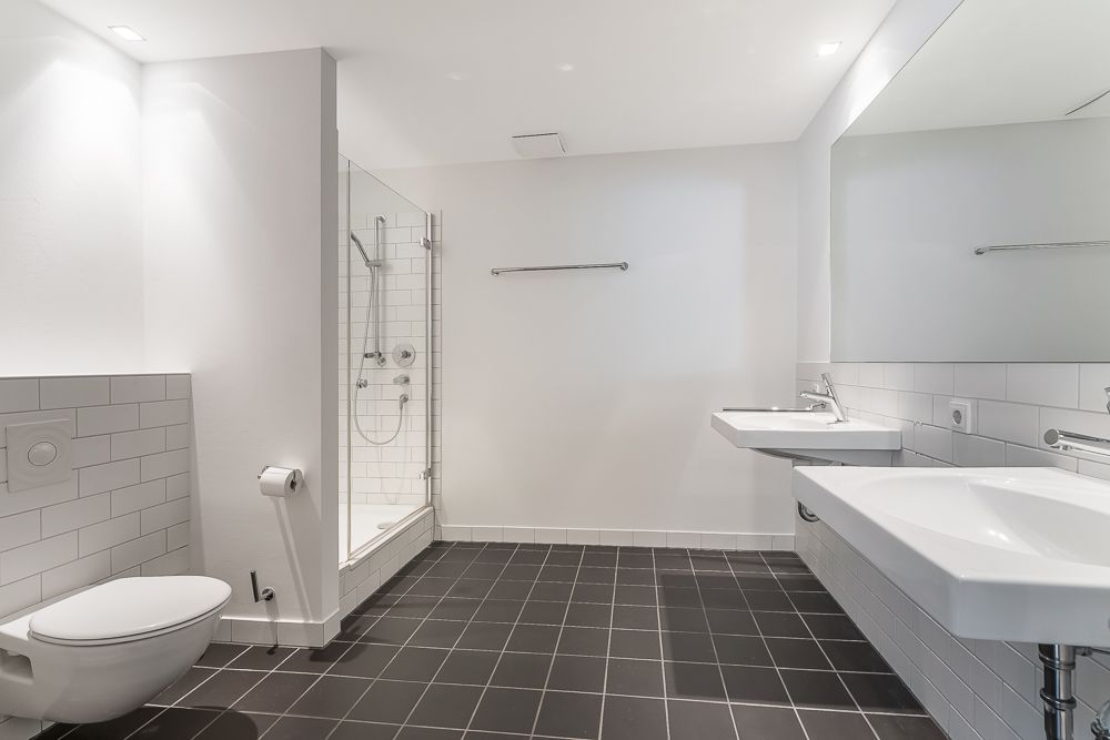 Modernes Bad mit Dusche, zwei Waschbecken und Toilette, weiße Wandfliesen und dunkler Boden.