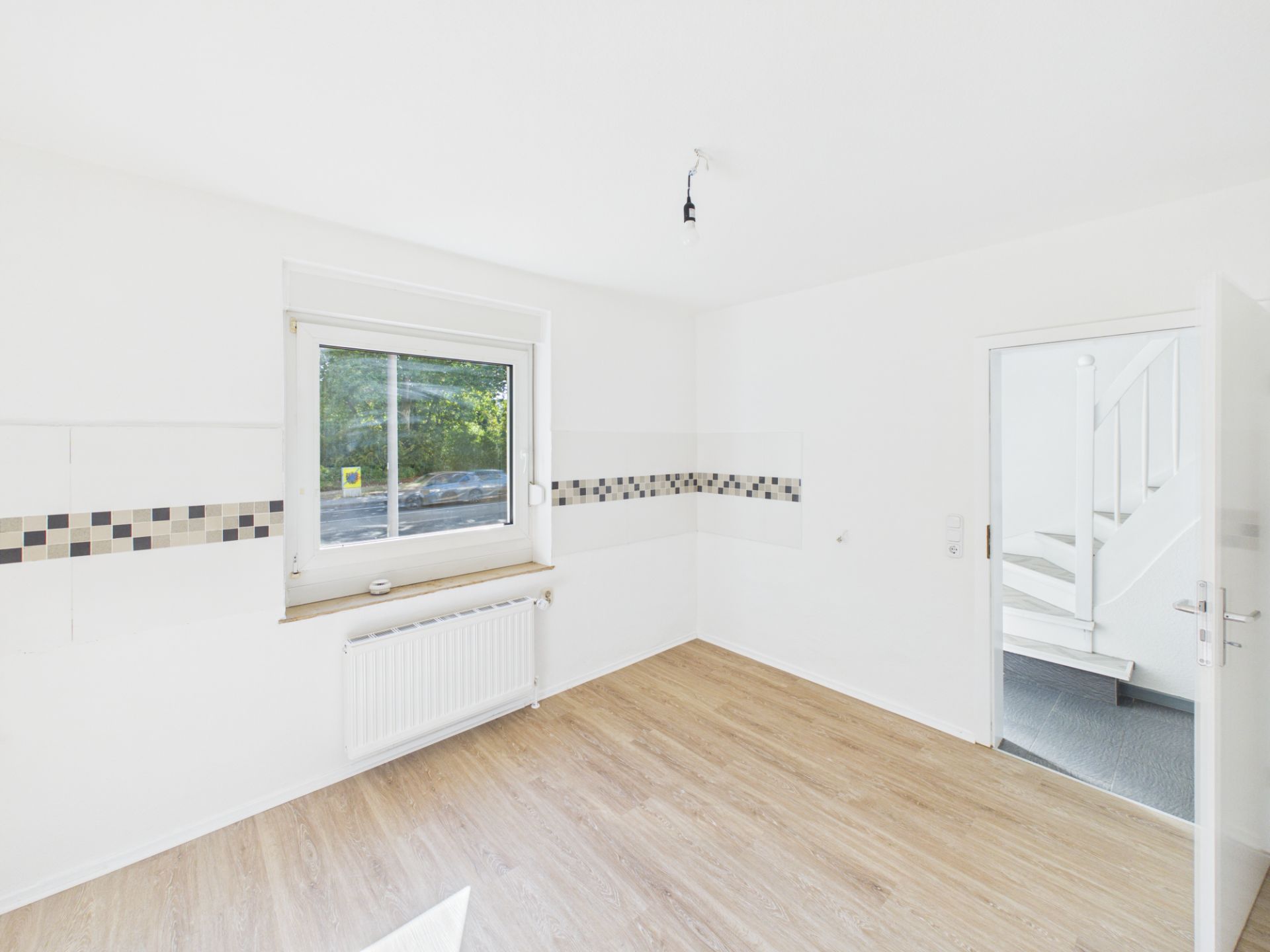 Heller Raum mit Fenster, Heizkörper und Holzboden, Tür führt zu einer Treppe.