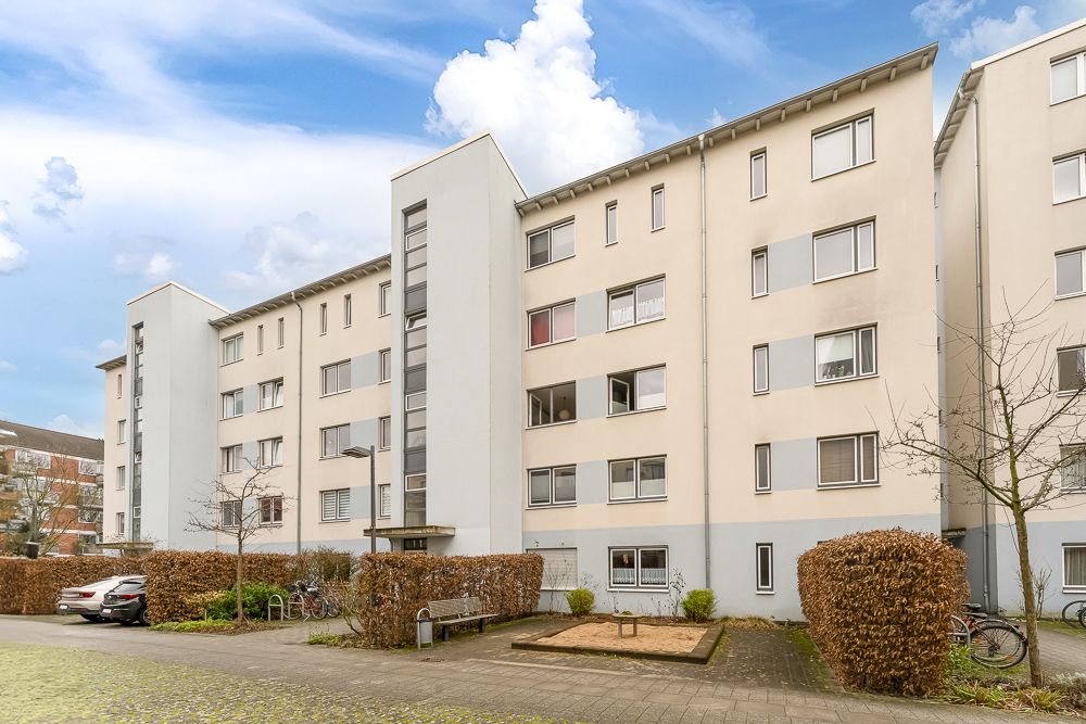Mehrstöckiges Wohngebäude mit mehreren Fenstern, Autos, Bänken und kleinen Bäumen davor.