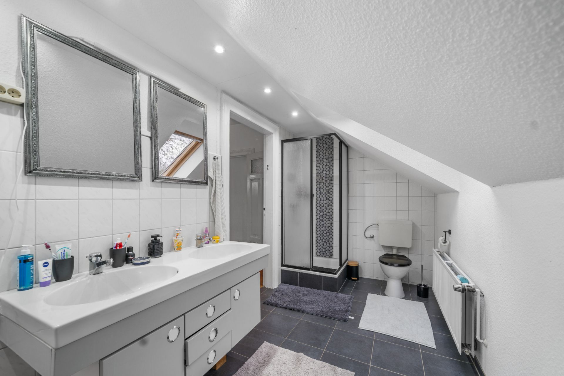 Modernes Badezimmer mit Doppelwaschbecken, Dusche, WC und Spiegel unter einer Dachschräge.
